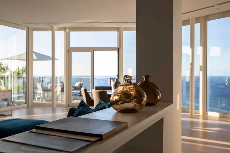 Modern hotel suite interior with ocean view through large windows, gold vases and notebook on teal counter.