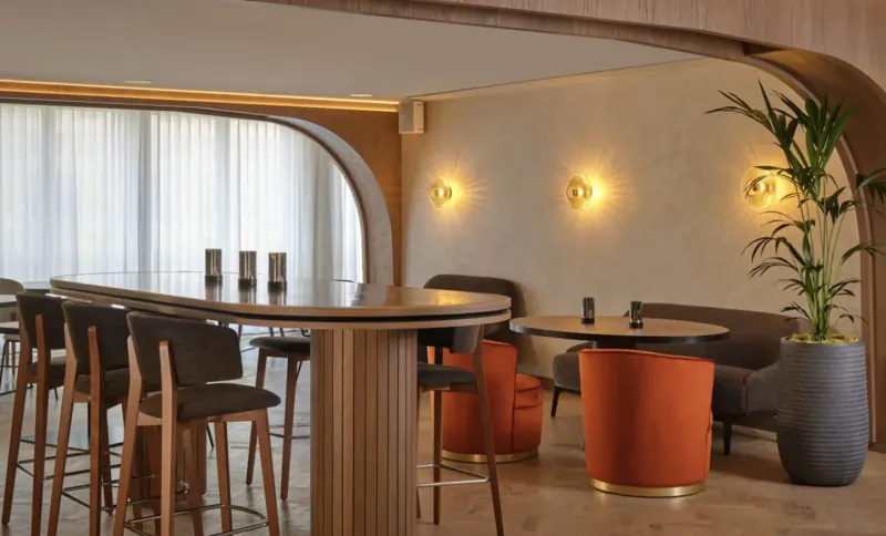 Modern hotel bar at The Reach at Piccadilly, Manchester, with curved wooden bar stools, orange seating, plants, and warm lighting.