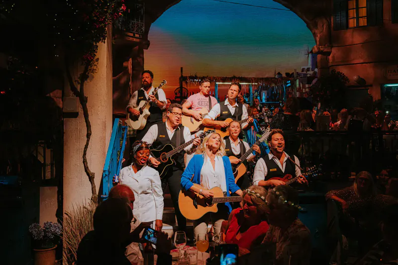 Cast of Mamma Mia! The Party performing energetically on guitars under a sunset balcony archway at The May Fair Hotel.