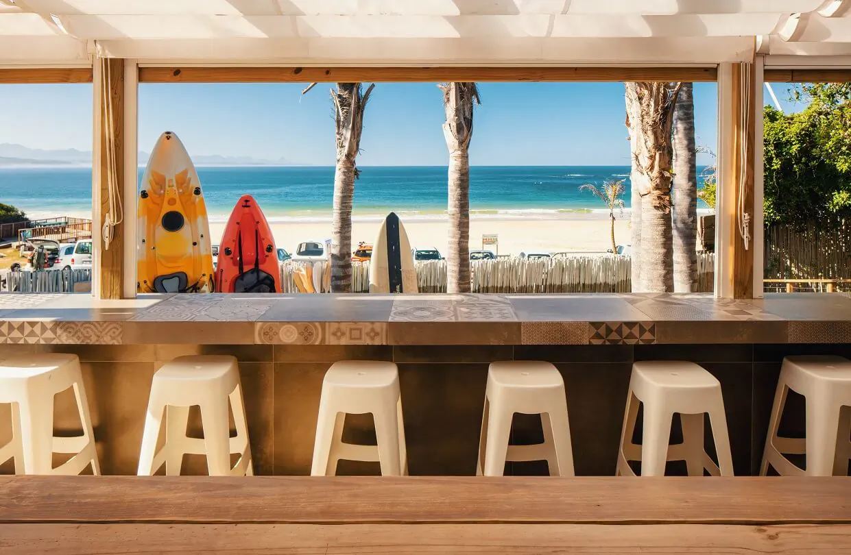 Beachfront bungalow bar with colorful surfboards, white stools, ocean view, and palm trees at The Bungalow Plett.