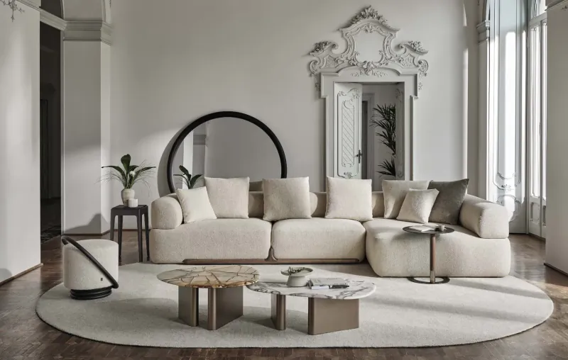 Modern white L-shaped sofa with cushions in luxurious room with ornate mirror, plants, marble tables, and round rug.