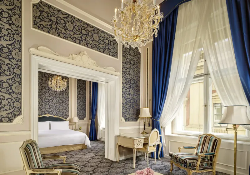 Opulent Hotel Imperial suite in Vienna with ornate gold chandelier, king bed, desk, blue velvet curtains, and brocade walls.