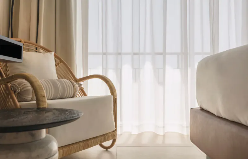 Cozy rattan armchair with cushions and side table beside bed in sunlit Hotel Riomar room with sheer curtains.