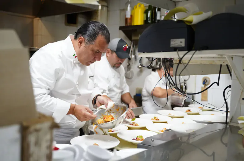 Chefs in white uniforms plating food on dishes in a busy professional kitchen for Italian event.