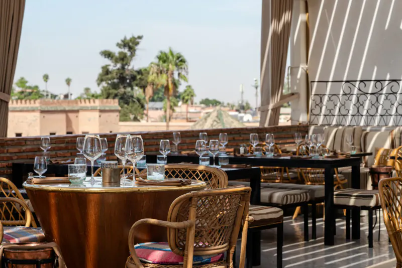 Rooftop restaurant in Marrakech with wicker chairs, round copper table, wine glasses, palm trees, and medina skyline.