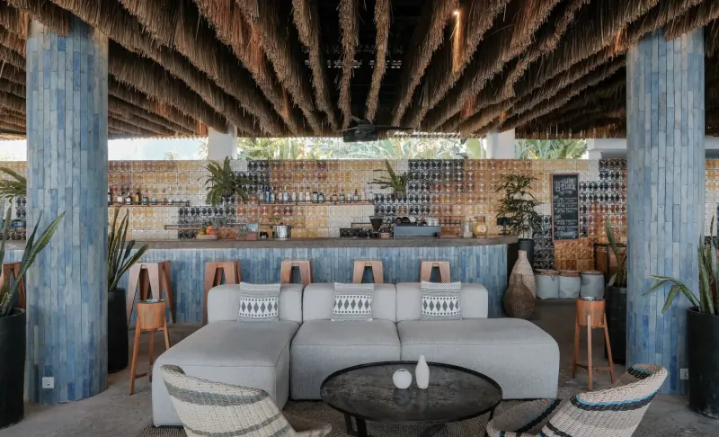 Open-air beach bar at Amber Lombok with thatched roof, blue pillars, wooden bar stools, potted plants, ocean views.