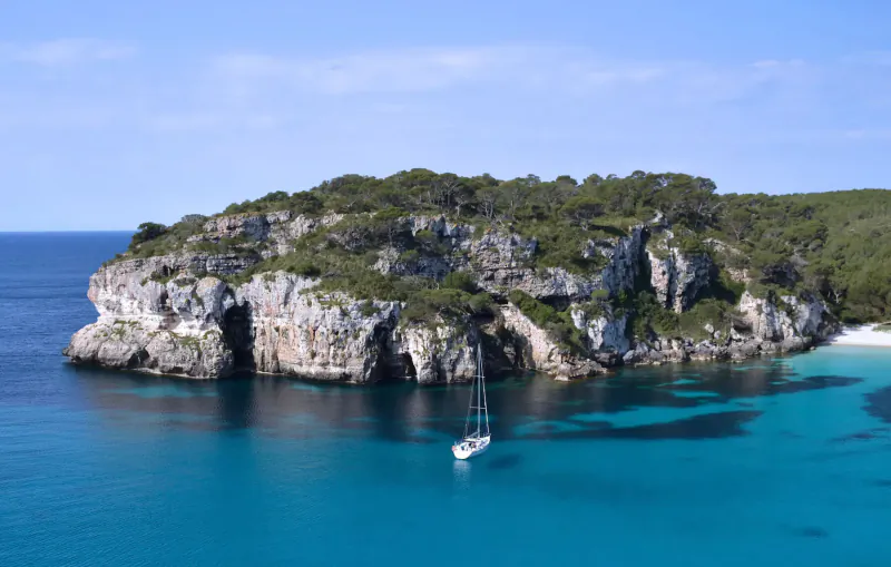 Sailboat anchored in turquoise cove with white sandy beach and lush green cliffs under blue sky