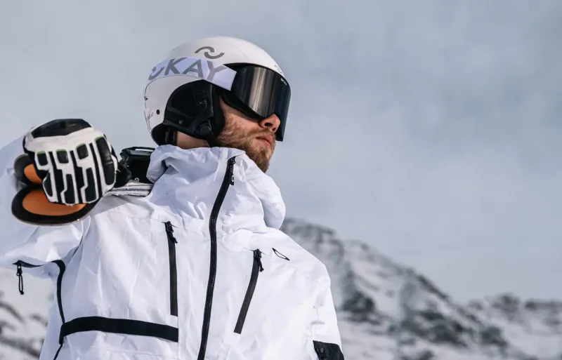 Bearded man in Zerokay helmet and white ski jacket holding skis, snowy mountains backdrop