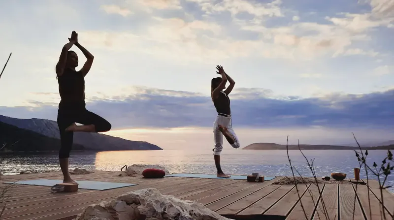 Two people in tree pose yoga on a dock at Maslina Resort's Pharomatiq Spa, sunset over lake and mountains.
