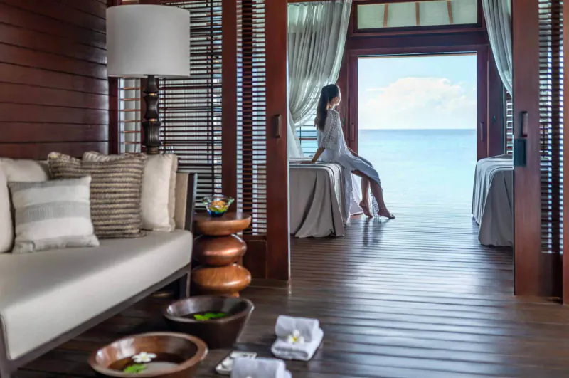 Woman in white robe sits on massage bed in luxurious overwater villa, gazing at ocean through open doors, foot baths and towels nearby