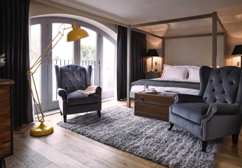 Elegant bedroom in Harper Norfolk with four-poster bed, yellow arched lamp, velvet armchairs, rug, and French doors to garden.
