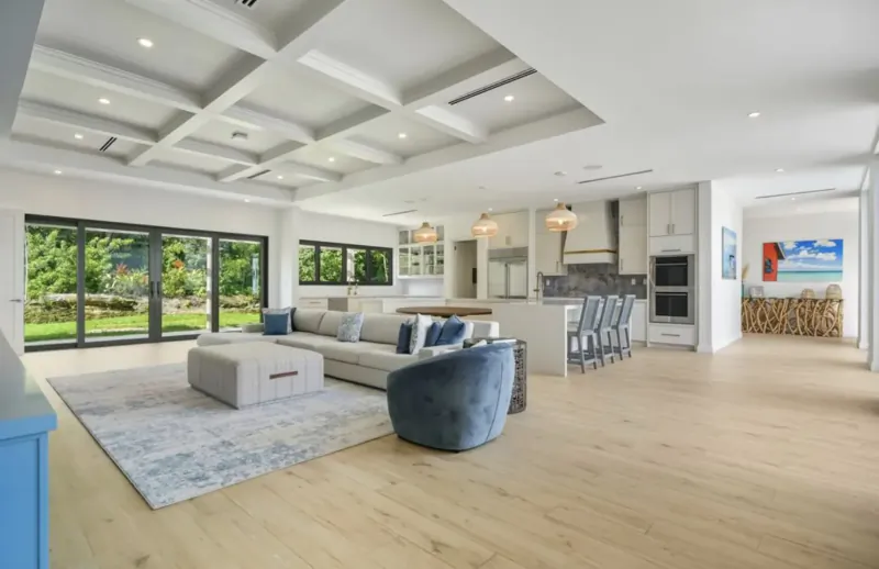 Spacious modern living room with white L-shaped sofa, blue velvet chair, open kitchen, and garden view through folding glass doors in Lyford Cay luxury home