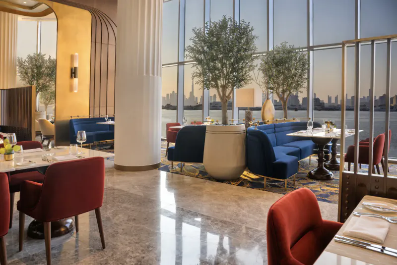 Elegant evening dining area at Address Grand Creek Harbour with blue velvet seating, olive trees, city skyline view, and set tables.