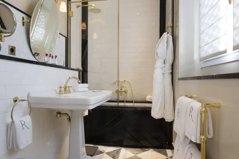 Elegant Relais Christine bathroom with white robe on gold hook, pedestal sink, black tub, gold fixtures, checkered floor.