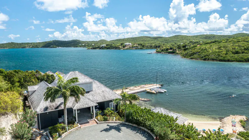 Aerial view of Rokuni luxury villa in Antigua with thatched roof, dock, turquoise bay, beach, and lush green hills under blue sky.