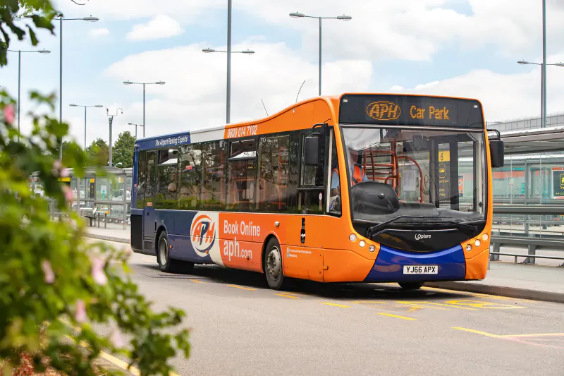 Orange and blue double-decker bus displaying 'API Car Park' at Gatwick Airport bus stop, with 'Book Online' ad, surrounded by flowers and streetlights.