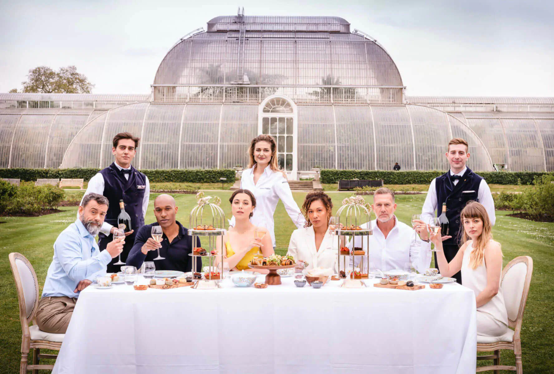 Group enjoying afternoon tea outdoors on white tablecloth, with waiters, before the grand glass Palm House greenhouse.