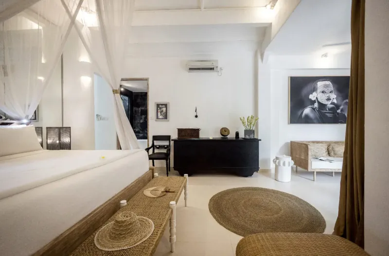Spacious white Bali villa bedroom with large bed, wooden desk, rattan hats, sheer mosquito net canopy, and black-and-white art.