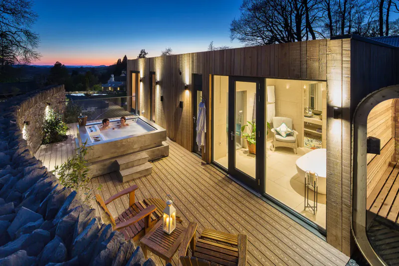 Twilight view of Gilpin Lake House: modern wooden building with hot tub on deck, person relaxing in tub, Lake District hills.