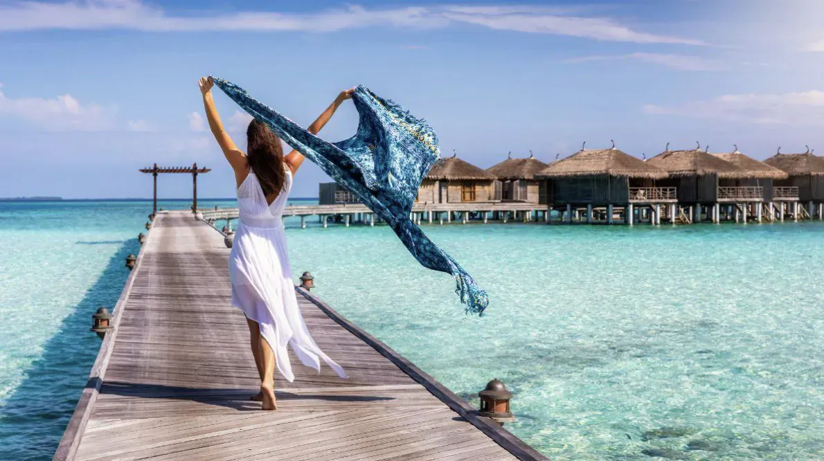 Woman in white dress walking on wooden pier over turquoise lagoon, holding flowing blue scarf, luxury overwater bungalows behind