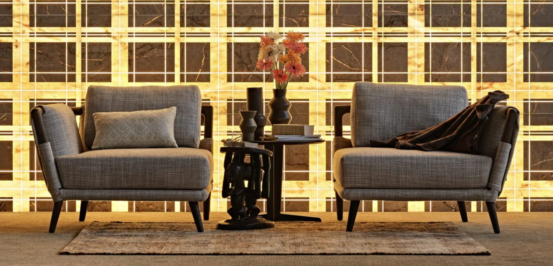 Two gray armchairs with pillows flank a round table holding orange flowers and sculptures, against glowing grid wall.