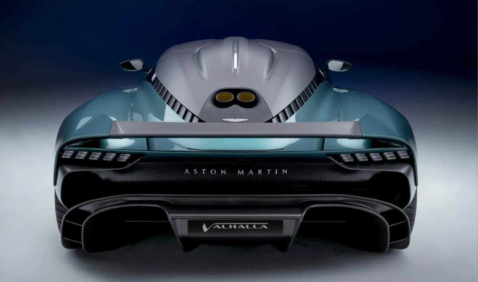 Rear view of teal Aston Martin Valhalla hybrid supercar with dual exhausts and Valkyrie badge on dark background.
