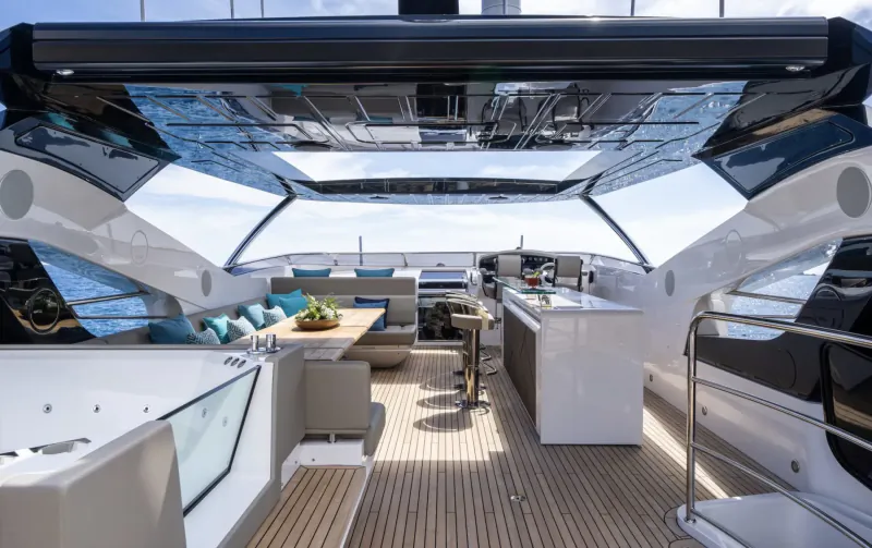 Luxury yacht deck with teak flooring, white lounge seating, turquoise cushions, bar stools, and galley kitchen under glass canopy.