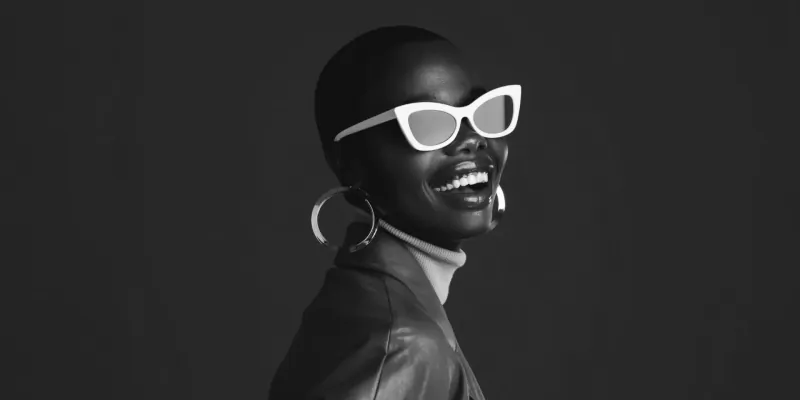 Smiling Black woman with bald head, white cat-eye sunglasses, large hoop earrings, black turtleneck and leather jacket, on black background