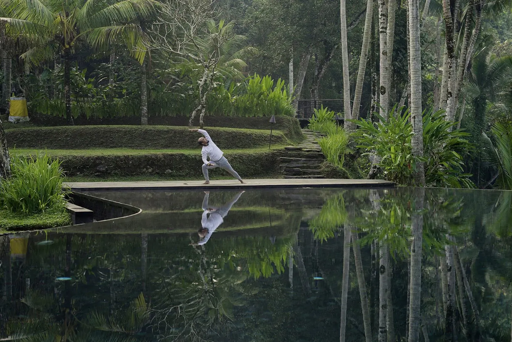 Woman in white performs yoga tree pose on wooden platform over reflective pool in lush Bali jungle villa.