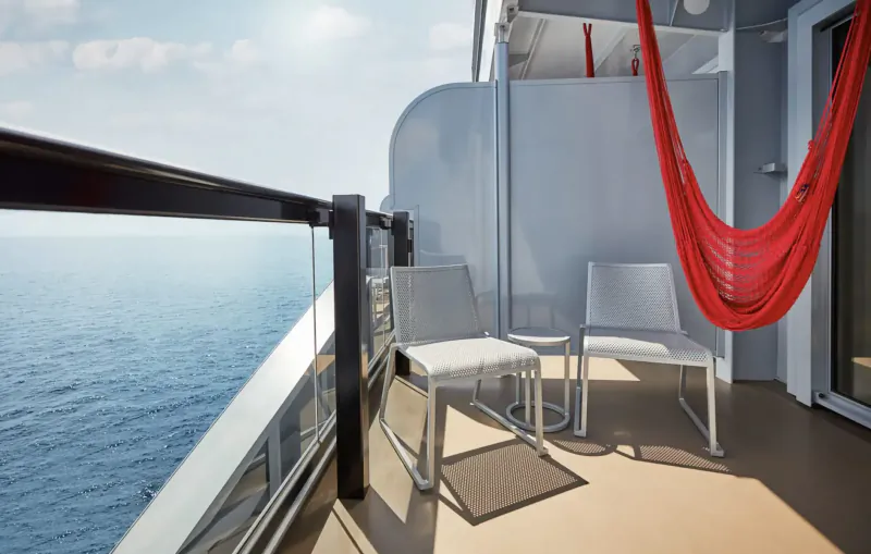 Cruise ship balcony with white chairs, red hammock, overlooking sunny ocean.