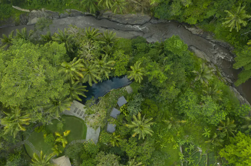 Aerial view of Kayumanis Ubud Bali resort in lush tropical forest, with blue pool, pavilions, lawns overlooking Ayung River.