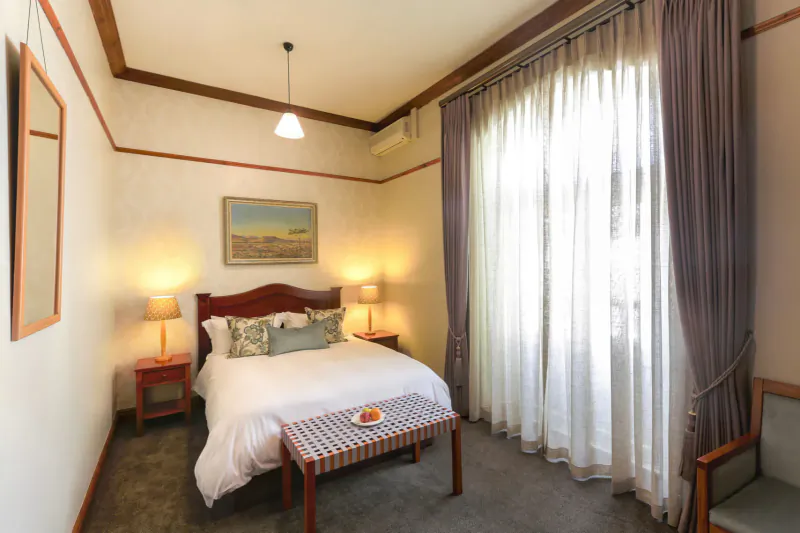 Cozy hotel room with wooden double bed, pillows, lamp tables, landscape painting, sheer curtains, and fruit on bench.