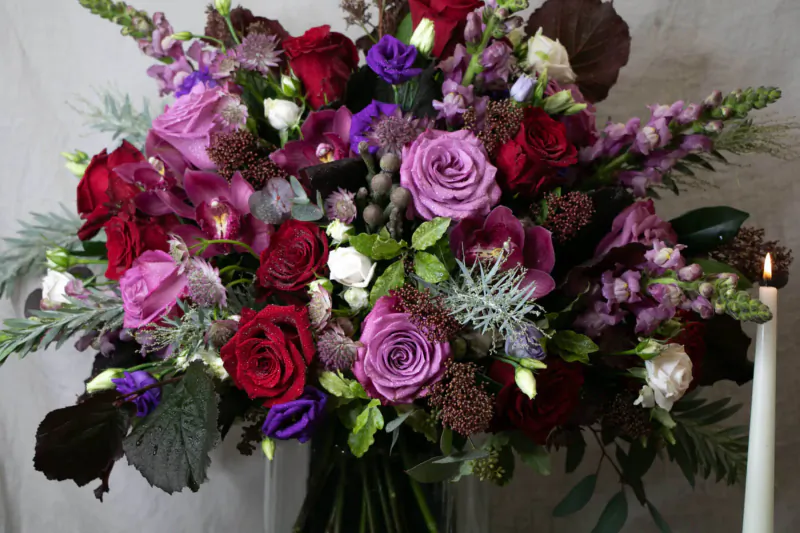 Vibrant bouquet of purple, red, and white roses with greenery and candles beside it
