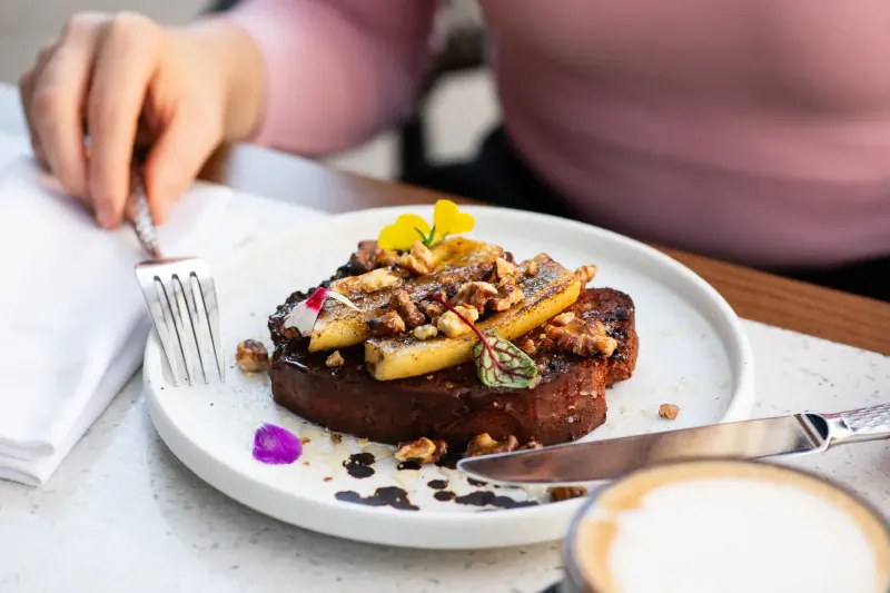Close-up of French toast topped with sliced bananas, walnuts, and viola flowers on a white plate, with coffee cup nearby