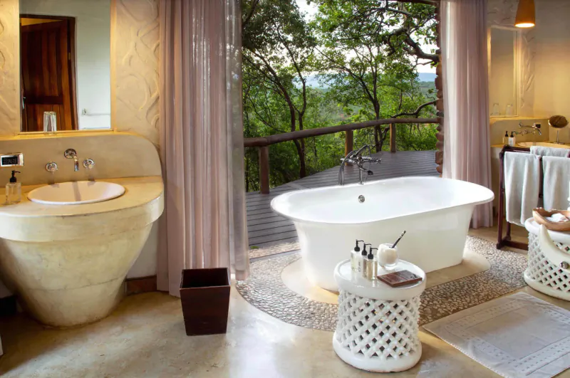 Luxurious bathroom at Madikwe Hills lodge with clawfoot tub, stone sink, and panoramic view of lush treetop deck.