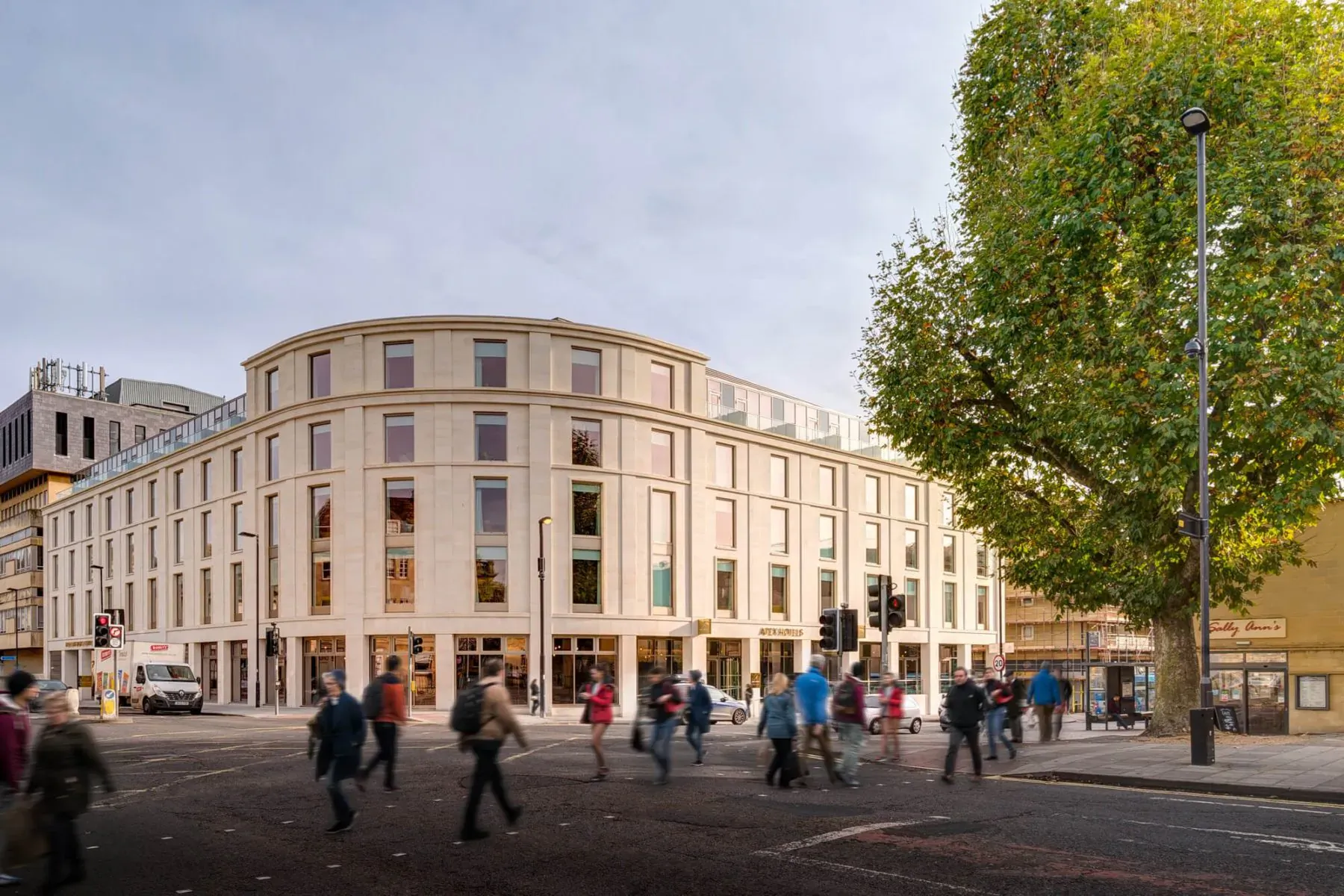 Curved Apex City of Bath Hotel facade with large windows, pedestrians crossing busy street, tall green tree beside it.