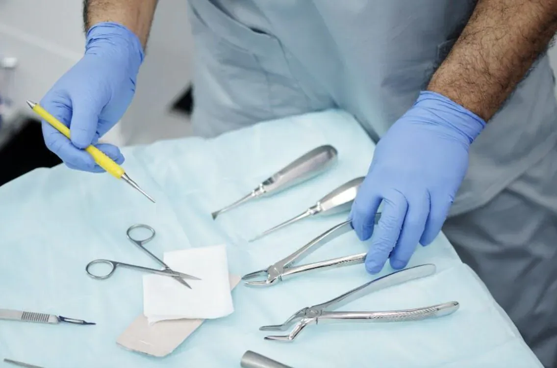 Surgeon in blue scrubs and gloves holding yellow pen over dental tools including retractors, forceps, scissors on blue drape
