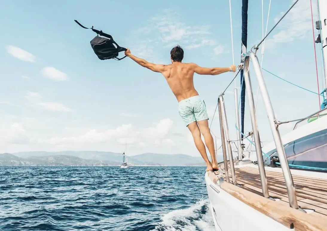 Shirtless man in light swim shorts leaps from yacht railing, holding black backpack, sea and mountains behind