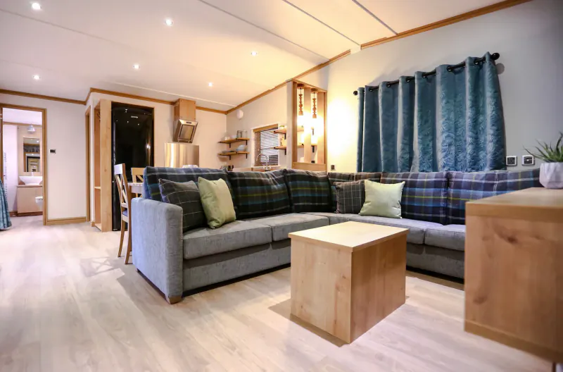 Cozy living room in Sandy Balls Holiday Village with L-shaped grey plaid sofa, wooden table, blue curtains, and plants.