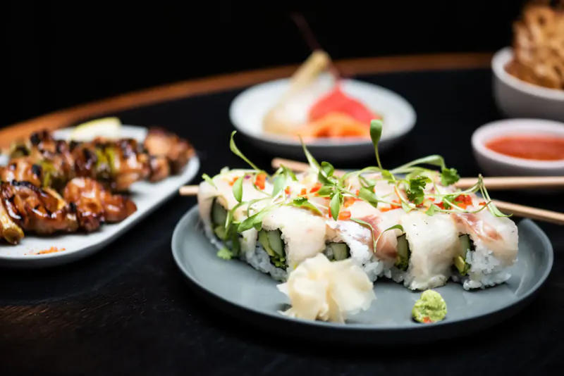 Assorted sushi rolls with microgreens and spicy mayo on plate, glazed chicken wings, pear salad, and chili sauce on black table.