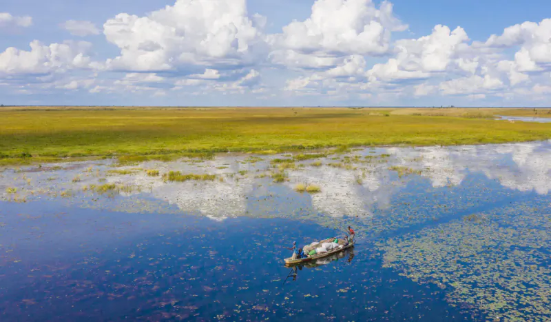 Aerial view of lone fisherman in wooden boat on Bangweulu Wetlands, surrounded by golden grasses, lily pads, and blue water under cloudy sky.