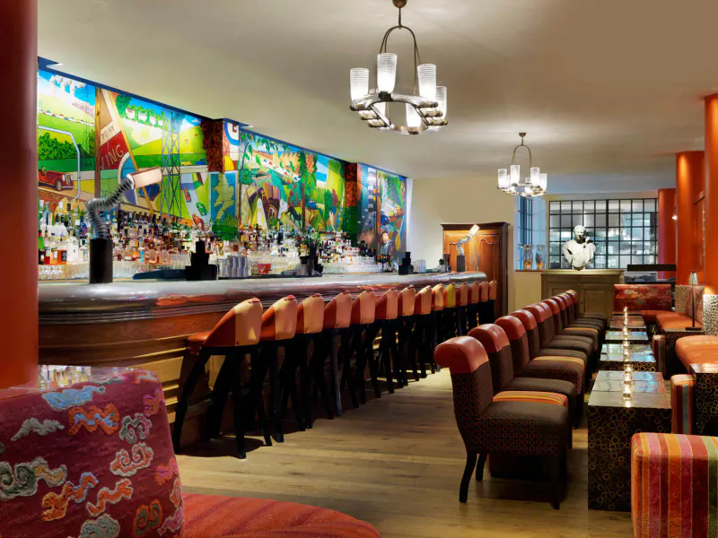 Chic Soho Hotel bar with colorful mural wall, wooden bar stocked with bottles, red stools, pendant lights, and lounge seating.