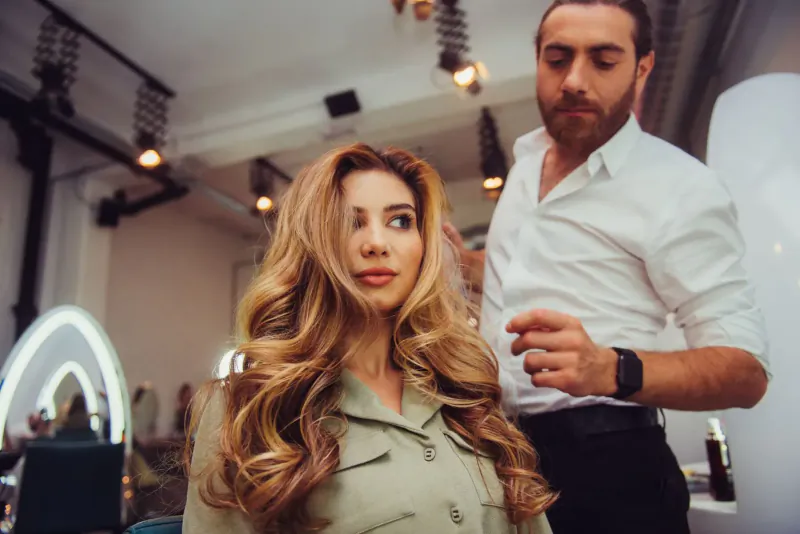 Bearded male stylist adjusting hair of woman with long wavy blonde hair in modern salon