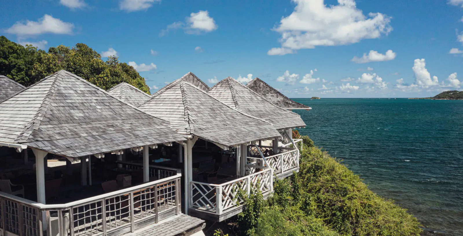 Rokuni in Antigua: thatched-roof bungalows with decks perched on a lush cliff overlooking the turquoise sea under blue skies.