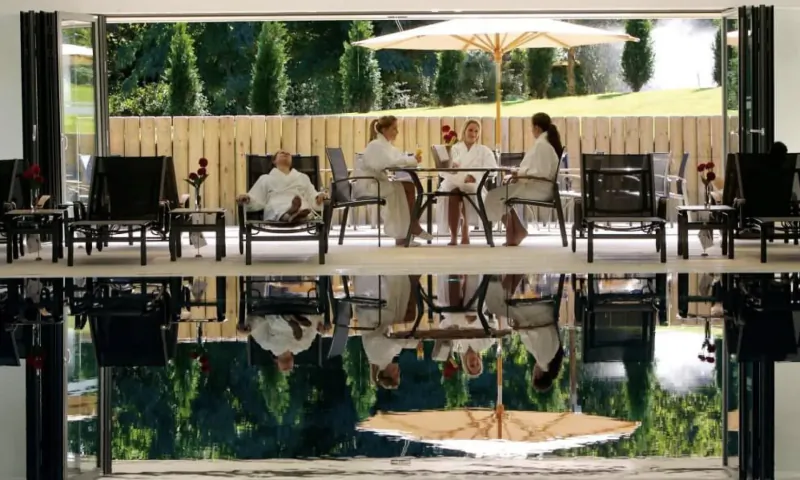 Group in white robes relaxing at poolside table in luxury spa, with golf course view and reflections in water.