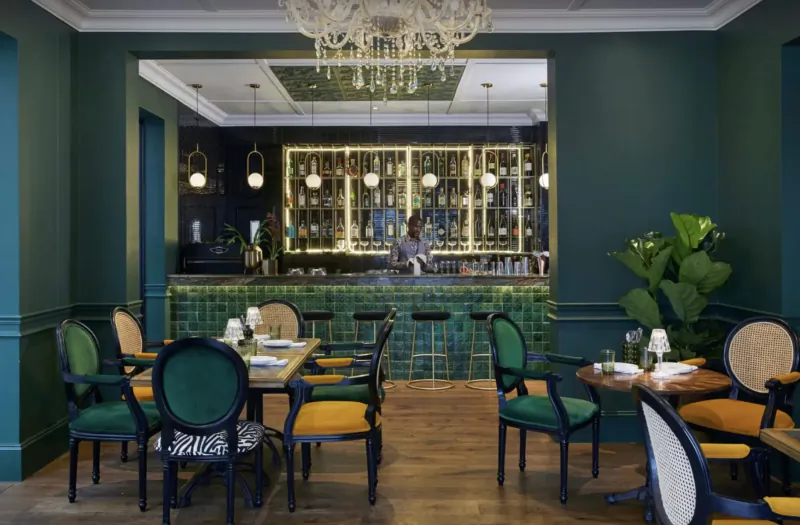 Elegant green-toned lounge at Cape Cadogan Boutique Hotel with ornate chandelier, bartender behind tiled bar, and colorful chairs at tables.