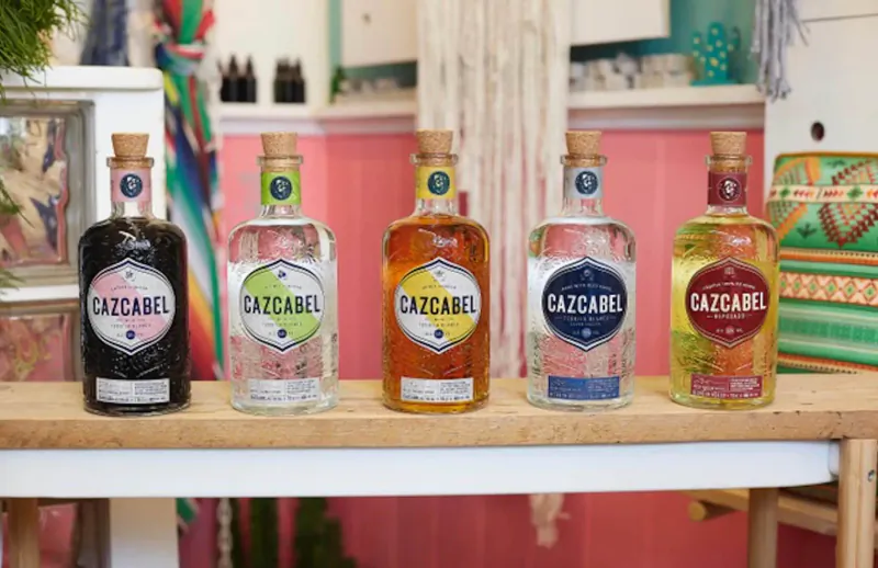 Five colorful Cazcabel tequila bottles on a wooden shelf in a boho pink shop setting.