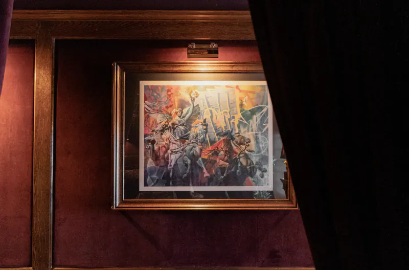 Framed abstract painting of vibrant figures in motion, lit in luxurious maroon gallery with wood frame and drapes.