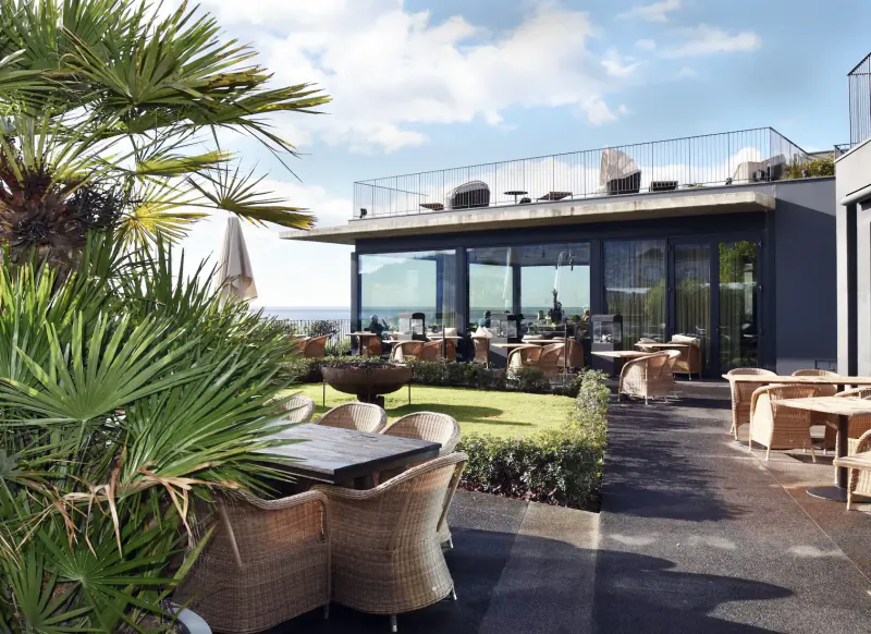 Outdoor terrace at Les Suites Hotel, The Cliff Bay, Funchal: wicker tables and chairs, palms, sea view through glass walls.