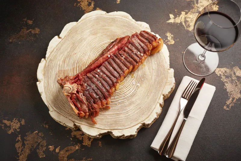 Seared ribeye steak on wooden board with fork and napkin, red wine glass beside, on gold-flecked table.